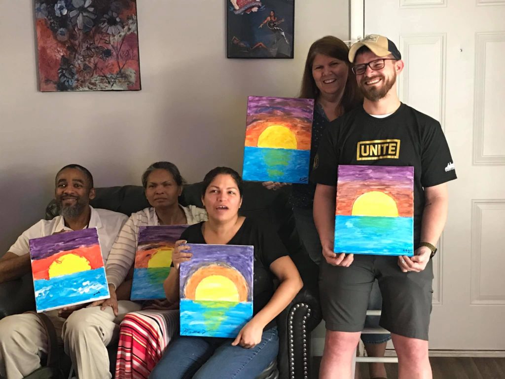 people holding finished paintings of sunset over the ocean